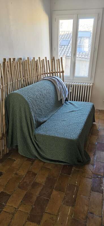 Une pièce rustique avec un canapé recouvert de vert, un dossier en bambou, un sol carrelé, un radiateur, une fenêtre avec vue sur les toits et une couverture grise drapée dessus.