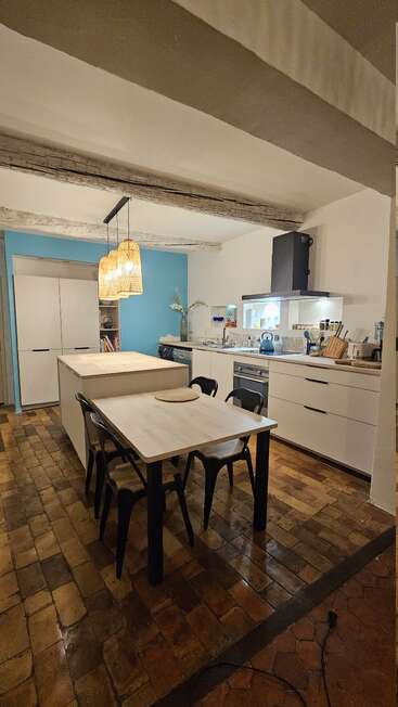 Cette image montre une cuisine confortable et moderne avec des poutres apparentes, un sol en briques, un mur d'accent bleu, un îlot, une table à manger, des chaises noires, des lampes suspendues et des comptoirs organisés.