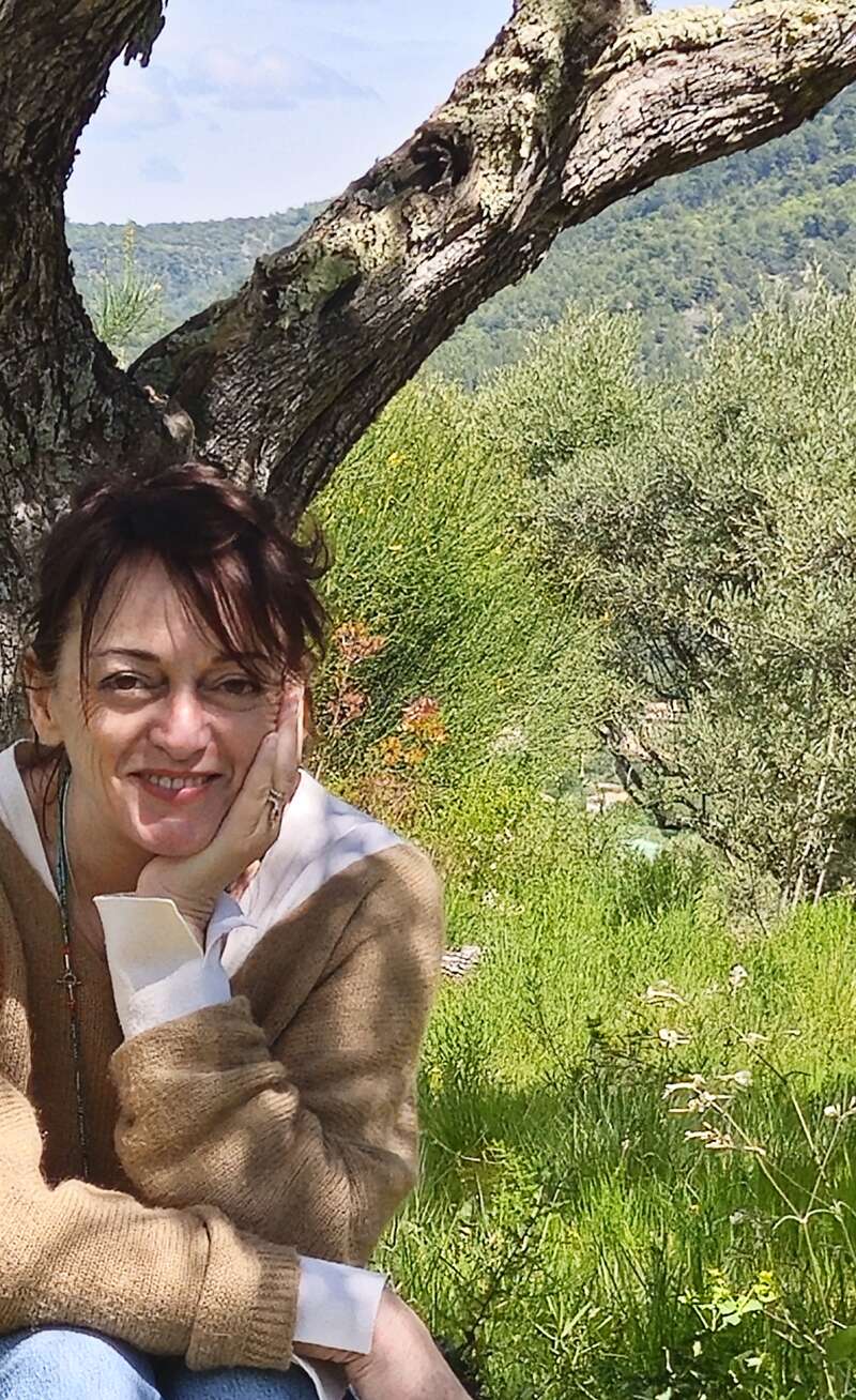 A woman sits under a tree with her hand resting on her face, smiling gently. Green grass and trees surround her, creating a peaceful outdoor scene.