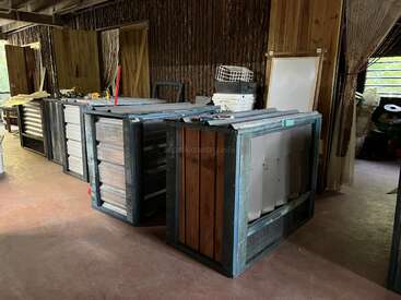 Um espaço de oficina com materiais de metal e madeira empilhados, ferramentas e suprimentos. O cômodo tem paredes de madeira, janelas abertas e uma atmosfera geralmente organizada, mas rústica.