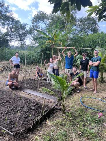 Un grupo de personas sonríen y posan juntas en un frondoso jardín, construyendo soportes para plantas y trabajando la tierra, rodeadas de vegetación y árboles bajo un cielo azul.