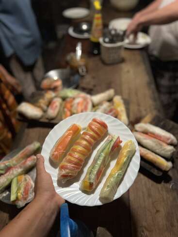 Una mano sostiene un plato de rollitos de primavera de verduras frescas. Se ven más rollitos de primavera e ingredientes sobre una mesa de madera, con gente preparando comida al fondo.