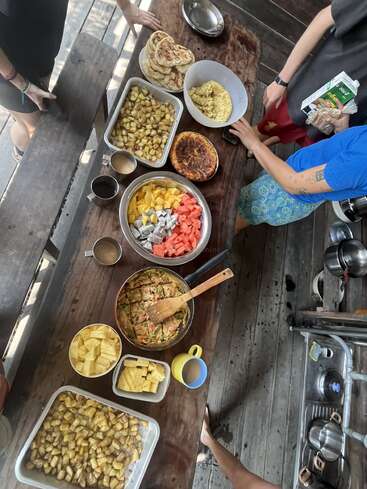 Una mesa de madera está puesta con patatas asadas, tortilla, fruta fresca, pan plano, café, y la gente reunida alrededor, preparándose para disfrutar juntos de una comida comunitaria.
