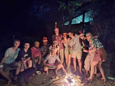 Un grupo de amigos se reúne alegremente alrededor de una hoguera por la noche, algunos con sombreros festivos, sosteniendo bebidas, sonriendo y disfrutando de la compañía de los demás en un acogedor entorno al aire libre.