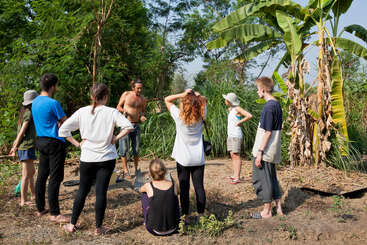 La imagen muestra a un grupo de personas de pie en un jardín, rodeadas de frondosos árboles, algunas de ellas vestidas con ropa informal y otras descalzas.