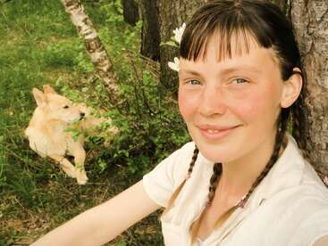 Une jeune femme aux cheveux tressés, une fleur glissée derrière l'oreille, sourit, assise en plein air adossée à un arbre, tandis qu'un chien au pelage clair se prélasse à ses côtés.