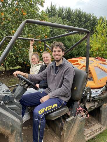 Trois amis sont assis, souriants, dans un véhicule tout-terrain entouré de verdure luxuriante et d'orangers, profitant d'une joyeuse aventure en plein air par une journée nuageuse.