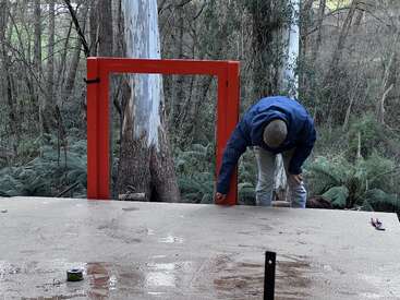 Une personne vêtue d'une veste bleue se penche près d'un cadre rectangulaire rouge, sur fond de forêt avec des arbres et des sous-bois verts.