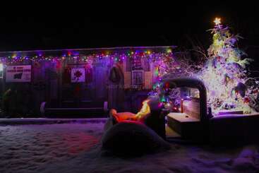 Um pátio nevado à noite apresenta uma árvore de Natal decorada, luzes coloridas em um edifício rústico e um caminhão velho coberto de neve e luzes festivas.