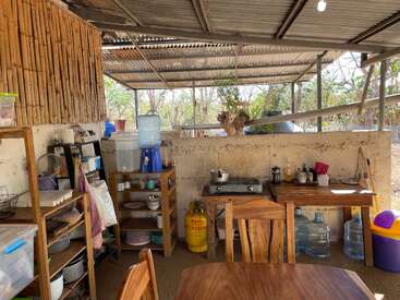 This image shows a rustic outdoor kitchen with wooden shelves, a table, a gas stove, water containers, dishware, bamboo walls, and sunlight streaming in.