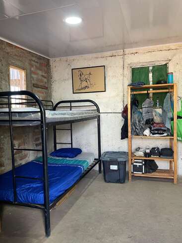 This image shows a simple room with a metal bunk bed, blue bedding, a wooden shelf with clothes, a storage bin, and a horse drawing on the wall.