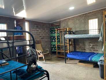 This image shows a simple, rustic dormitory room with bunk beds, concrete walls, a wooden rocking chair, open shelving, and minimal decorations. Natural light enters.