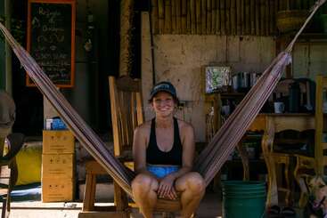 A smiling woman sits comfortably in a hammock, wearing a cap and casual clothes. She is surrounded by wooden furniture, books, and a green bucket, outdoors.