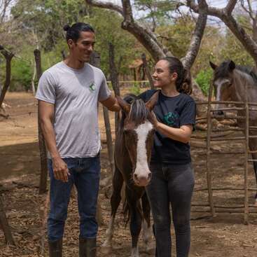 A man and woman stand outdoors with a young horse, smiling at each other. Another horse is in the background. The setting appears to be a farm.