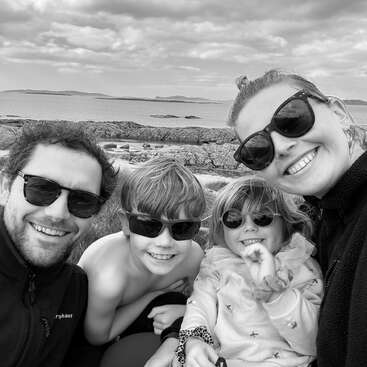 Une famille souriante de quatre personnes portant des lunettes de soleil pose pour un selfie en plein air sur un rivage rocheux, avec l'océan et des collines lointaines sous un ciel nuageux. Photo en noir et blanc.