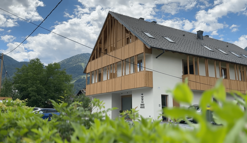 Ein modernes Haus im Chalet-Stil mit Holzvertäfelung und großen Fenstern. Umgeben von viel Grün liegt es unter einem blauen Himmel mit vereinzelten Wolken und Bergen.