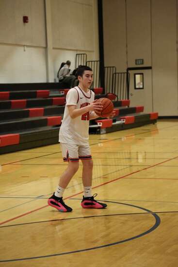 Un jeune basketteur en uniforme blanc s'entraîne sur le terrain d'un gymnase. Les gradins sont en grande partie vides, seules quelques personnes regardent en arrière-plan.