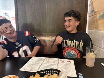 Deux garçons sont assis à la table d'un restaurant, souriant et riant. L'un porte un maillot des Patriots et mange des frites ; l'autre porte une chemise d'été Nike. Menus visibles.