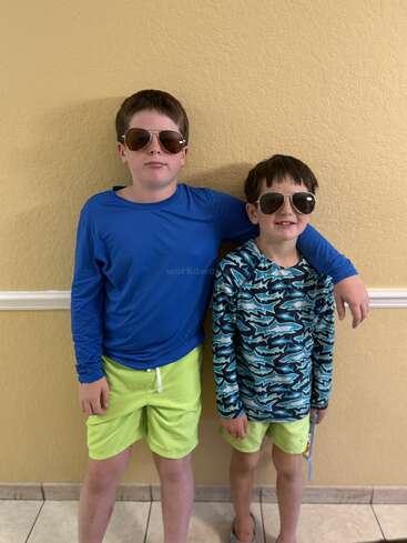 Deux garçons portant des lunettes de soleil et des shorts de bain assortis se tiennent contre un mur jaune. Le plus âgé a le bras autour du plus jeune, tous deux souriant.
