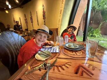 Deux enfants souriants, coiffés de chapeaux, sont assis à une table de restaurant décorée, en train de déguster un repas. Derrière eux se trouvent des décorations murales colorées et une grande fenêtre avec de la verdure.