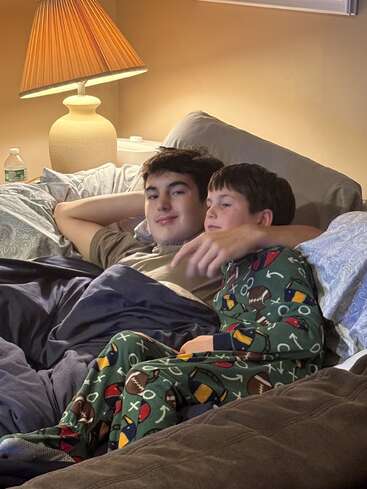 Deux garçons se détendent ensemble dans leur lit sous des couvertures douillettes, l'un d'eux portant un pyjama de football. Une lampe chaude brille derrière eux, créant une atmosphère paisible et réconfortante.