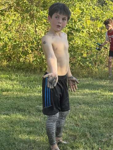 Un garçon boueux et torse nu se tient sur l'herbe, montrant des mains et des jambes sales. Un autre enfant à l'arrière-plan tient une canne à pêche près de buissons verts et de la lumière du soleil.