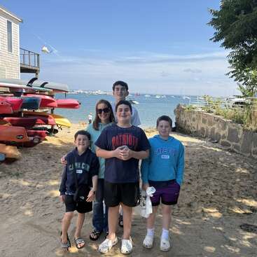 A group of five people stands on a sandy beach near colorful kayaks, smiling at the camera. The ocean and docked boats are visible behind them.