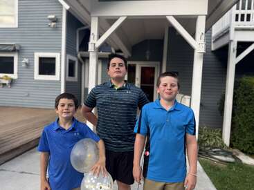 Trois garçons se tiennent ensemble devant une maison. Deux d'entre eux portent des chemises bleues, un autre des rayures, et le garçon de gauche tient des ballons transparents.