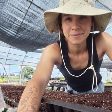 Una mujer con sombrero de ala ancha sonríe en un invernadero, cuidando bandejas de tierra. Lleva un detallado tatuaje en el brazo y parece contenta.