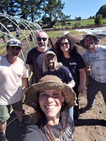 Sechs lächelnde Leute machen an einem sonnigen Tag draußen ein Selfie. Sie stehen auf einem Bauernhof mit Beeten, Metallgestellen und Bäumen im Hintergrund.