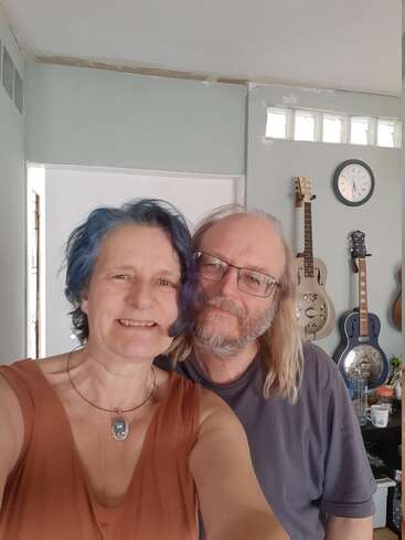 The image shows a couple posing for a selfie in a room with guitars on the wall, a clock, and a white door behind them.