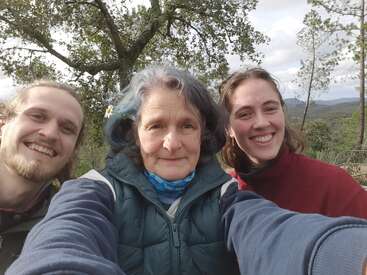 Três pessoas estão tirando uma selfie ao ar livre. Elas estão sorrindo, curtindo a natureza e parecem felizes. Árvores e colinas formam um fundo bonito e tranquilo atrás delas.