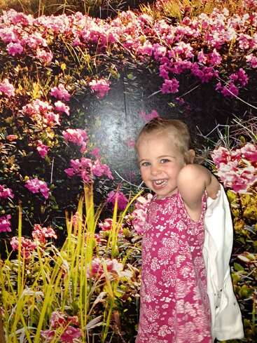 Ein lächelndes kleines Mädchen in einem rosafarbenen Blumenkleid steht freudig vor einem farbenfrohen Feld mit blühenden rosa Blumen und grünem Gras.