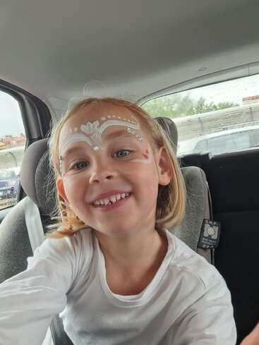 Ein junges, lächelndes Kind mit Gesichtsbemalung und Strasssteinen sitzt in einem Autositz und trägt ein weißes Hemd. Im Hintergrund sind ein Fenster und ein anderes Fahrzeug zu sehen.