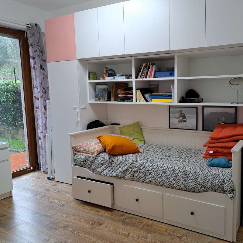 Das Bild zeigt ein gemütliches Zimmer mit einer Liege, Regalen und Schränken, einem Fenster mit Blick nach draußen und einer Tür, die in einen anderen Raum führt.