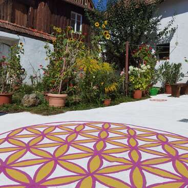 Ein farbenfroher Innenhof mit Topfpflanzen, Sonnenblumen und blühenden Blumen. Der Boden ist mit einem großen geometrischen Mandala-Muster in Gelb und Rosa verziert, das dem Ganzen einen künstlerischen Charme verleiht.