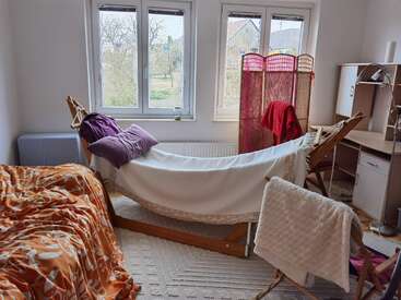 Cette chambre confortable est équipée d'un hamac avec des coussins, d'un lit à motifs orange, d'un bureau, d'une chaise, d'une cloison et de grandes fenêtres donnant sur les arbres, créant ainsi une atmosphère lumineuse et paisible.
