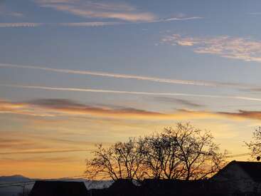 The image shows a beautiful sunset sky with streaks of clouds, silhouetted trees, rooftops, and mountains in the distance, creating a peaceful evening scene.