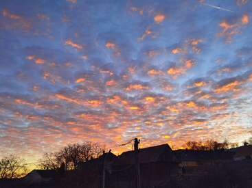 A stunning sunset sky filled with scattered, glowing orange clouds above the silhouette of houses, trees, and power lines, creating a peaceful village evening atmosphere.