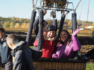 Two smiling women sit in a hot air balloon basket, waving, ready for takeoff. Others stand nearby. The background shows colorful autumn trees and clear skies.