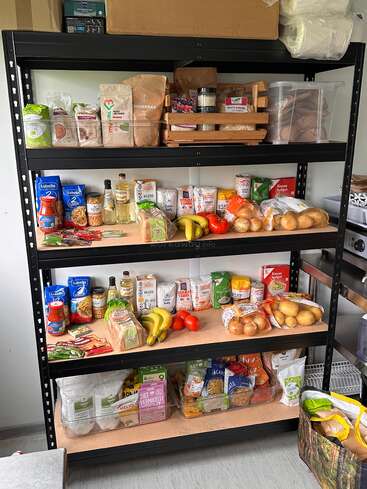 Ein Metallregal ist mit verschiedenen Lebensmitteln gefüllt: Nudeln, Konserven, Getreide, Zwiebeln, Bananen, Tomaten und verpackte Produkte sind übersichtlich angeordnet und leicht zugänglich.