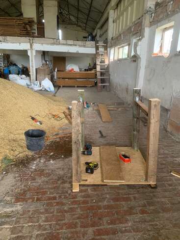A partially constructed wooden structure stands in a large, rustic workshop. Tools, sawdust, wooden planks, and materials are scattered around. There’s a loft and ladder visible.