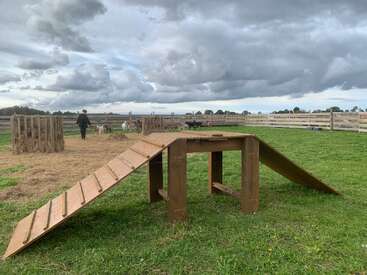 A wooden ramp structure stands on green grass in a fenced farm area. Goats and a person are seen in the background under a cloudy sky.