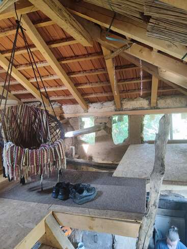 Ein gemütliches Loft mit einem rustikalen Holzdach, einem in eine bunte Decke gehüllten Hängesessel, Schuhen auf einer Matte und einer Person, die darunter arbeitet. Natürliches Licht strömt herein.