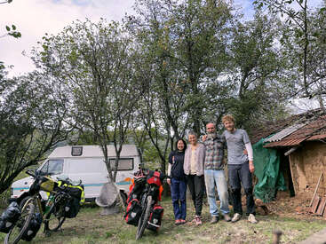 Vier Menschen stehen lächelnd zusammen im Freien, umgeben von Bäumen. Zwei beladene Fahrräder sind in der Nähe geparkt, neben einem Wohnmobil und einem rustikalen Schuppen mit Blechdach.