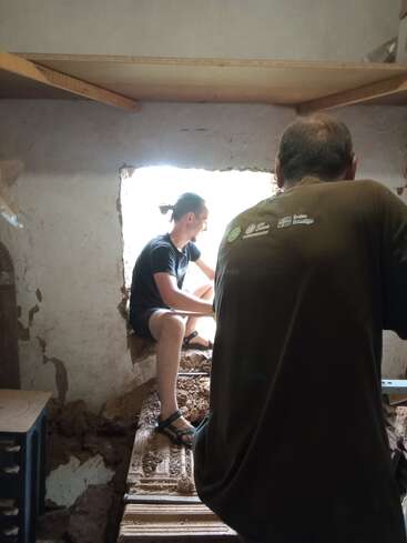 Zwei Menschen arbeiten in einem Raum mit rauen, unfertigen Wänden. Einer sitzt in einer Fensteröffnung, durch die das Sonnenlicht einfällt, während der andere in der Nähe steht, teilweise verdeckt.