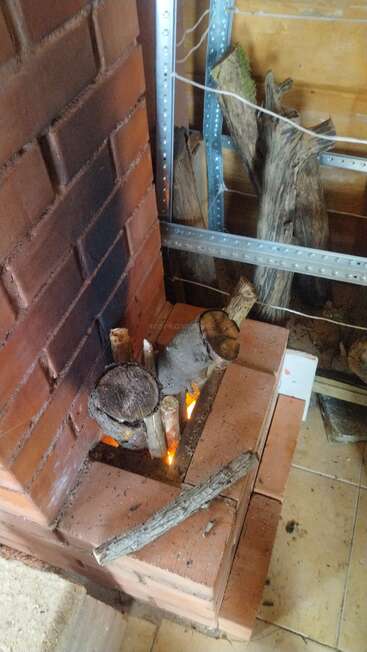 En una estufa de ladrillo arde un pequeño fuego de leña. Sobre los ladrillos hay leña cortada y ramitas, y cerca, apoyada contra una estructura metálica, hay más leña apilada.