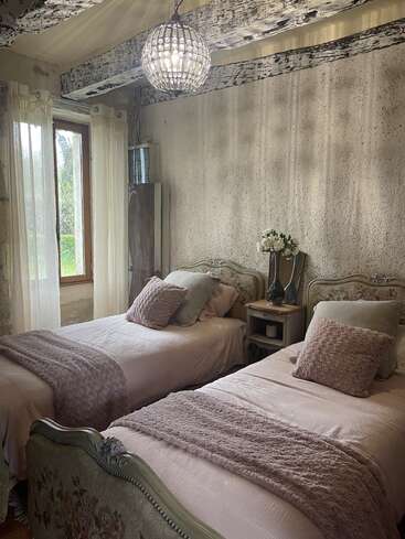 Este quarto aconchegante tem duas camas de solteiro vintage com roupa de cama em tons pastéis, mantas texturizadas, vigas de madeira rústicas, cortinas transparentes, detalhes florais, um lustre e uma luz natural suave.