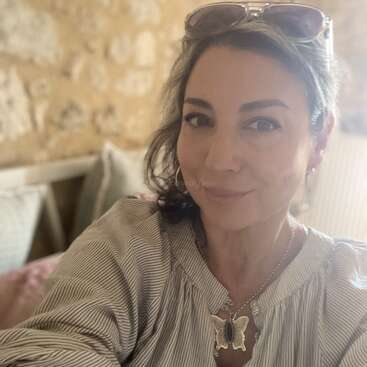 Uma mulher com óculos escuros na cabeça sorri para uma selfie. Ela está vestindo uma blusa listrada e um colar de borboleta. O fundo tem uma iluminação suave e aconchegante.