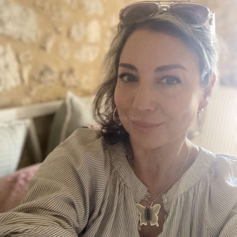 Uma mulher com óculos escuros na cabeça sorri para uma selfie. Ela está vestindo uma blusa listrada e um colar de borboleta. O fundo tem uma iluminação suave e aconchegante.